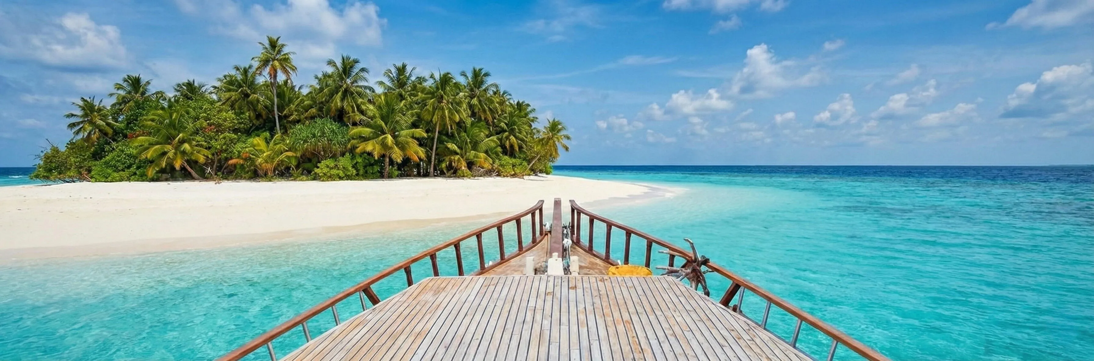 A small uninhabited palm island with a white sandbar, seen from the bow of a wooden boat on turquoise water