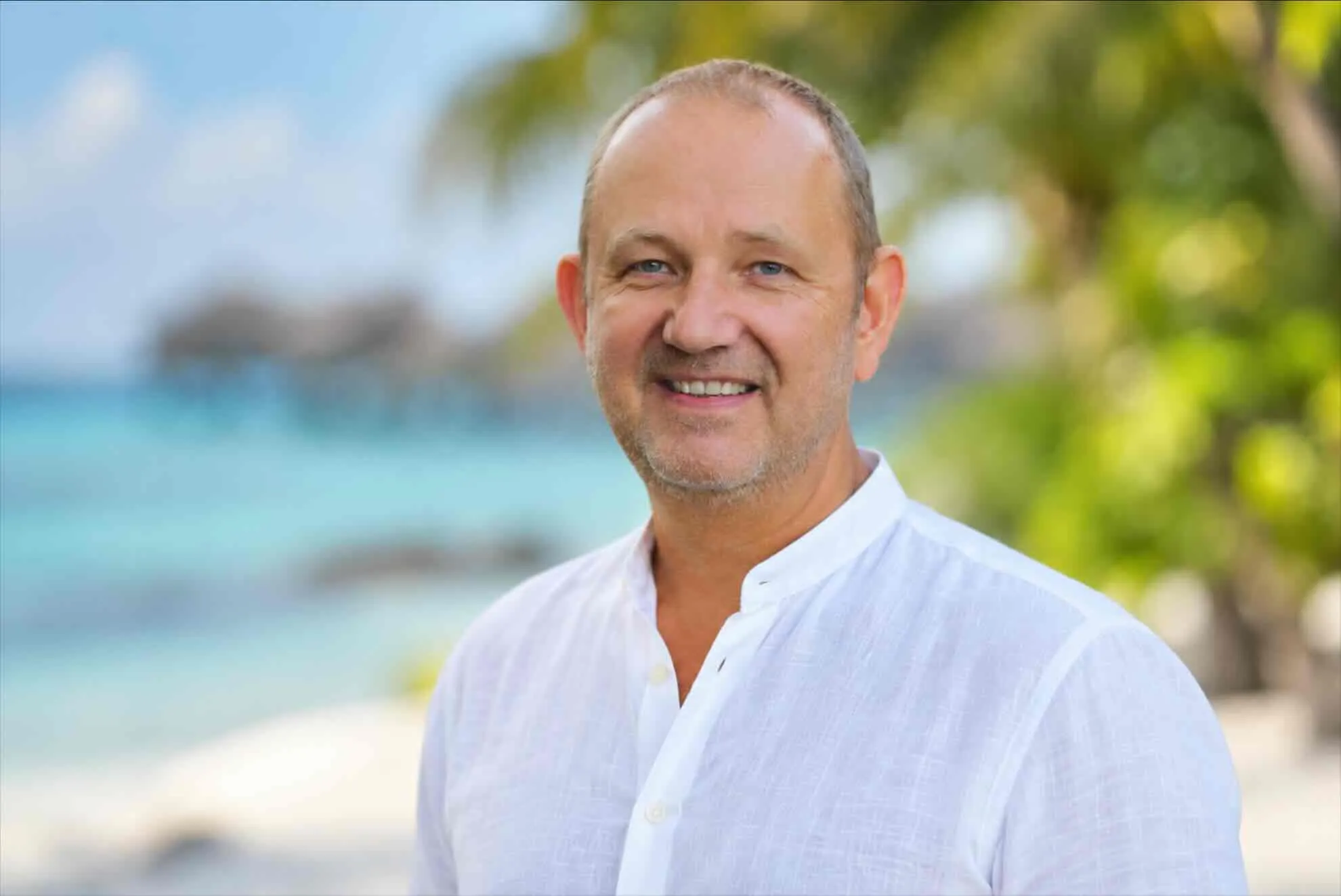 Portrait of Toddy, smiling, wearing a white shirt in front of a beach